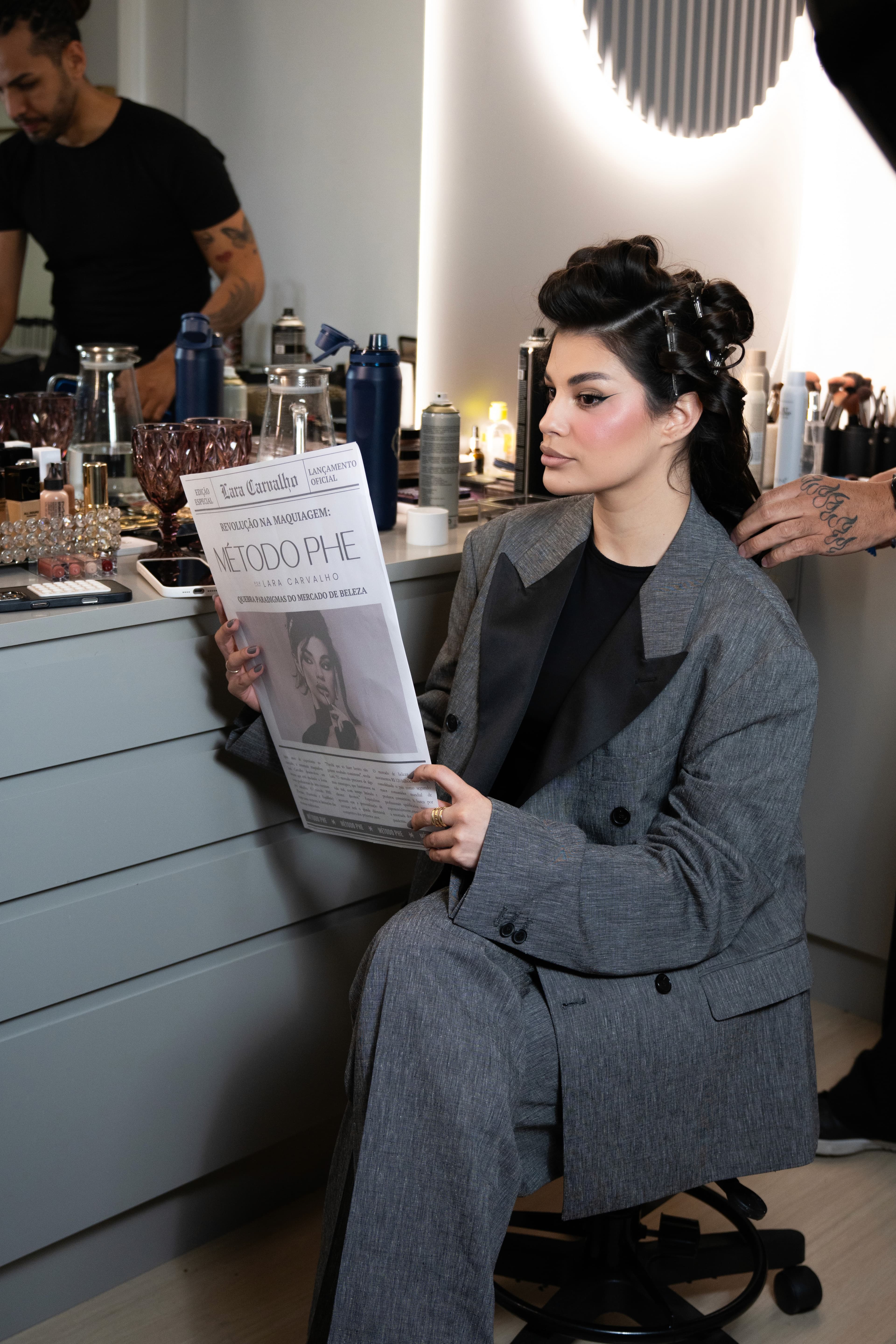 Lara sentada no camarim com o jornal, equipe ao fundo