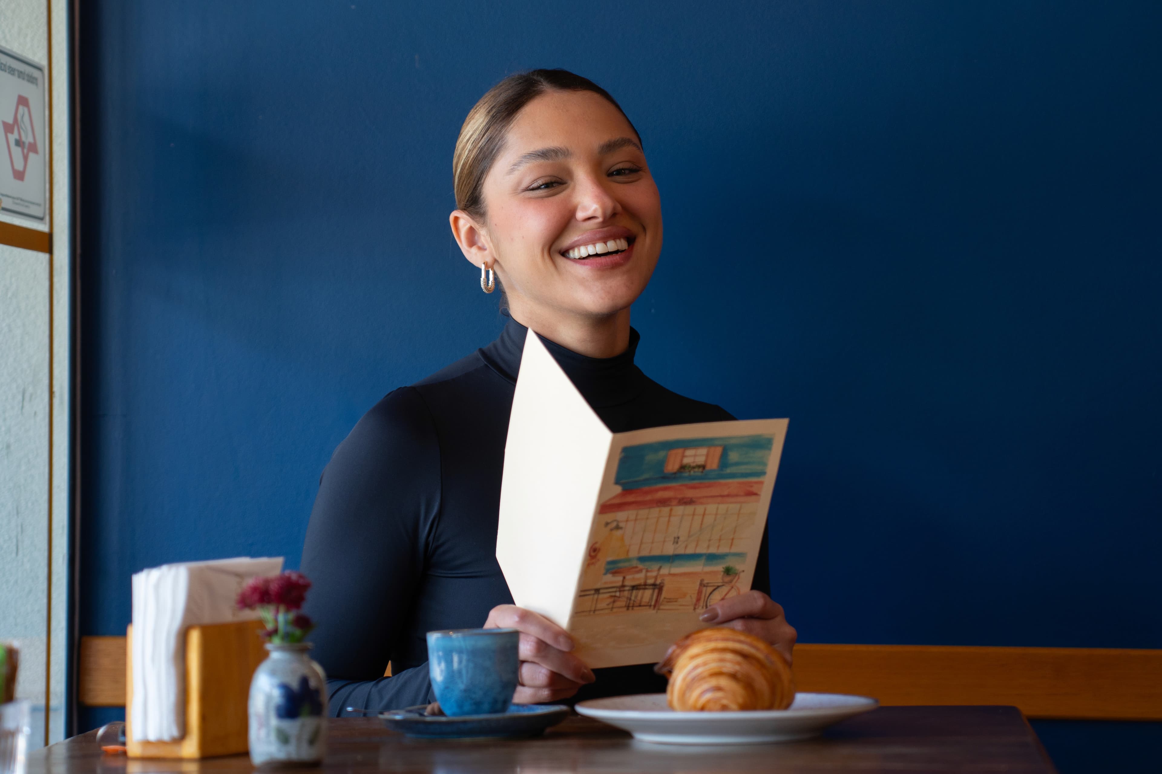 Cliente sorrindo com o cardápio, croissant e café na mesa